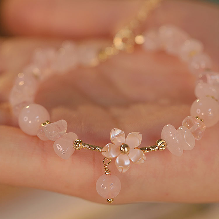 A delicate bracelet with pale pink stone beads, a small flower charm, and gold-tone metal accents, resting on a person's hand.